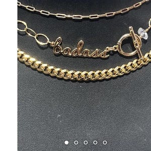 May include: Three gold chains with a charm that says "badass". The charm is attached to a toggle clasp. The chains are layered on top of each other.