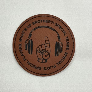 May include: Brown leather coaster with a black outline and the text "What's Up Brother?! Special Teams, Special Plays" with a black silhouette of a hand giving a thumbs up wearing headphones.
