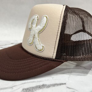 May include: A brown and beige trucker hat with a white embroidered letter "K" on the front. The hat has a brown mesh back.