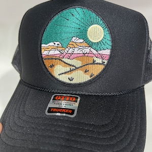 May include: Black trucker hat with a circular embroidered patch featuring a mountain landscape with a sun and a teal sky. The patch has the text "OTTO WORLD FAMOUS FOAM FRONT OS/FM TRUCKER".