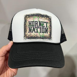May include: White and black trucker hat with a black patch that says "Hornet Nation" in white letters. The patch has a green outline of the state of Texas with a floral pattern.