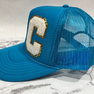May include: A turquoise blue trucker hat with a white felt letter "C" outlined in gold glitter. The hat has a mesh back.