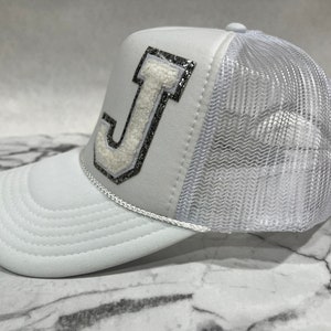 May include: White trucker hat with a white felt letter J with silver glitter accents on the front. The hat has a white mesh back.