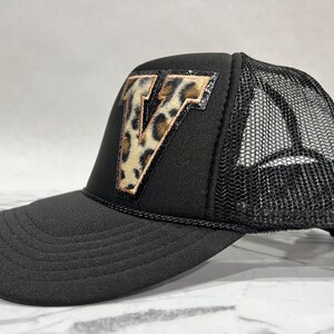 May include: Black trucker hat with a black mesh back and a leopard print letter "V" patch on the front.