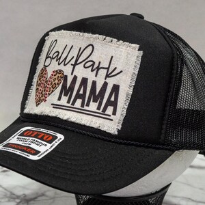 May include: Black trucker hat with a white patch that says "Ball Park Mama" with a leopard print heart. The hat has a black mesh back.