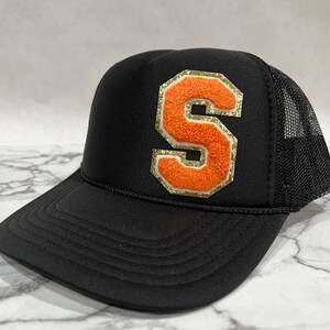 May include: A black trucker hat with a large orange felt letter "S" with gold trim on the front. The hat has a black mesh back.