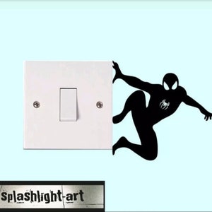 Könnte beinhalten: Eine weiße Lichtschalterabdeckung mit einer schwarzen Silhouette von Spider-Man, der auf der rechten Seite klettert. Die Spider-Man-Figur hat ein Spinnenemblem auf der Brust. Der Text "splashlight-art" befindet sich unten.