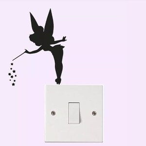 May include: Black silhouette of a fairy with a wand and stars, positioned above a white light switch. The fairy is depicted with large wings and a flowing dress. The light switch is rectangular with a single toggle.