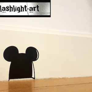 May include: A black silhouette of a mouse head with large ears, positioned near the floor against a white wall. The image includes the text "splashlight-art" in a rectangular frame at the top.