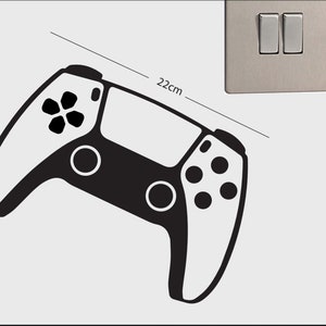 Op de afbeelding: Zwart-witte muursticker van een gamecontroller, 22 cm breed. De sticker toont een moderne gamecontroller, met knopdetails en een strak ontwerp. Een lichtschakelaar is zichtbaar op de achtergrond.