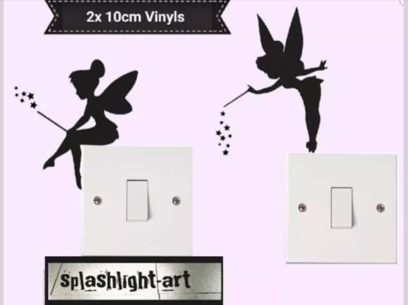 May include: Two black fairy vinyl decals, each 10cm in size, are shown on a light purple background. One fairy is seated, and the other is standing. Both are holding wands with star accents. The decals are placed above white light switch covers.