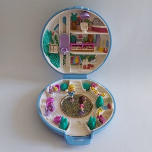 Op de afbeelding: Een vintage blauw en wit Polly Pocket speelset, open om een miniatuurwereld te onthullen. De speelset heeft twee kleine figuren, een tuin en een interieur met meerdere niveaus met meubels en accessoires. Een nostalgisch speelgoed.