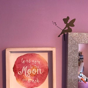 May include: A framed print with the words "love you to the Moon and Back" over a watercolor design. A decorative fairy silhouette sits on a glittery frame. The background is a solid lavender color.
