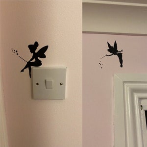 May include: Black silhouette fairy wall decals. One fairy is seated on a light switch, holding a wand with stars. The other fairy is flying near a white door frame, also holding a wand with stars. The decals are on a pink wall.