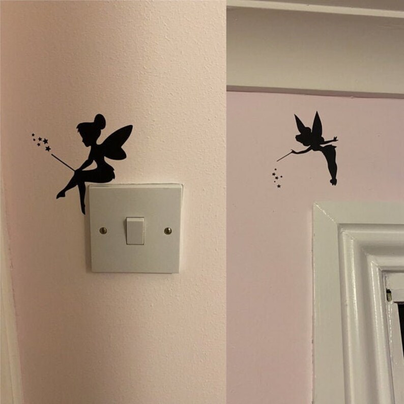 May include: Black silhouette fairy wall decals. One fairy is seated on a light switch, holding a wand with stars. The other fairy is flying near a white door frame, also holding a wand with stars. The decals are on a pink wall.