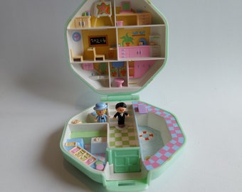 Może przedstawiać: Zabytkowy, miętowy zestaw Polly Pocket, otwarty, aby odsłonić szkołę i basen. Szkoła ma tablicę z napisem "2+2=4". Dwie miniaturowe figurki stoją w strefie basenu.