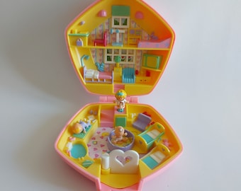 May include: A vintage, open, pink and yellow miniature dollhouse with multiple rooms and small figurines. The dollhouse features a heart-shaped bed, a baby in a tub, and other furniture. The interior is decorated with wallpaper and small details.