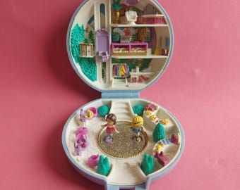 Może przedstawiać: Otwarta, niebiesko-biała, zabytkowa zabawka Polly Pocket. Wnętrze ukazuje miniaturowy świat z dwiema postaciami, okrągłym parkietem tanecznym i dwupiętrowym budynkiem z meblami i ubraniami. Na różowym tle.