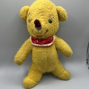 May include: A vintage yellow plush bear with a red bib. The bear has embroidered facial features, including blue eyes and a dark red nose. The plush toy has a worn appearance, suggesting it is an antique or well-loved item.