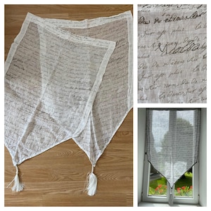 May include: Two white sheer curtains with a brown script pattern and tassels. The curtains are hung in a window with a view of green foliage.