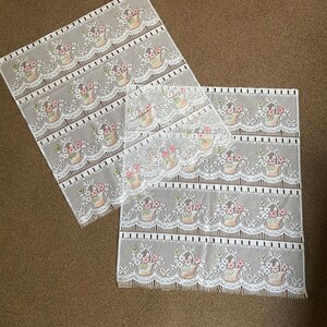 May include: White lace panels with a repeating pattern of flower baskets. The baskets are tan with pink and white flowers. The lace has a scalloped edge. The panels are on a brown surface.