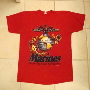 May include: Red t-shirt with a graphic of the US Marine Corps emblem. The emblem features a globe, an eagle, and an anchor. The text "Marines" and "The few. The proud. The Marines." is printed below the emblem.