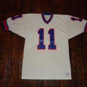 May include: Off-white football jersey with the number 11 in blue and red trim. The jersey has a V-neck collar and red and blue accents on the sleeves. The number 1 is also on the sleeves.