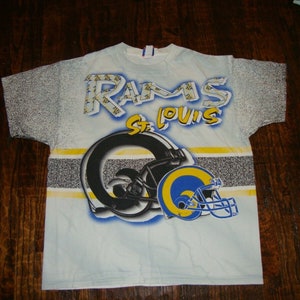 May include: A white t-shirt with a blue, yellow, and black graphic print of the St. Louis Rams logo. The shirt has a faded, distressed look.