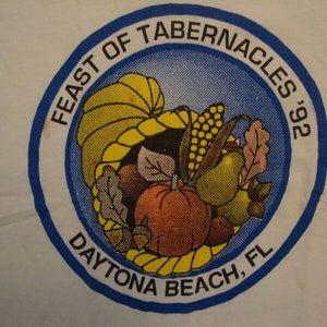 Może przedstawiać: Biała koszulka z okrągłą grafiką dla 'Feast of Tabernacles '92' w Daytona Beach, FL. Projekt zawiera róg obfitości wypełniony jesiennymi produktami, takimi jak dynie, kukurydza i gruszki, w niebiesko-białej obwódce.