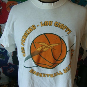May include: White t-shirt featuring a basketball graphic with the text "SAM PERKINS - LOU CIOFFI BASKETBALL ACADEMY" in a circular design. The basketball is orange with a yellow and green design. The shirt has short sleeves.