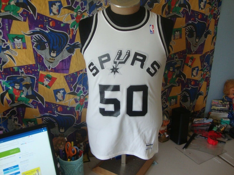 May include: White basketball jersey with black and silver trim. The jersey has the word "SPURS" in black letters with a silver star and a silver basketball logo. The number "50" is printed in black on the front of the jersey.