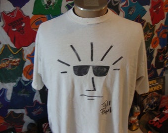 May include: White t-shirt featuring a black graphic of a face with sunglasses and radiating lines. The shirt has a crew neck and short sleeves. The artist's signature is visible below the graphic.