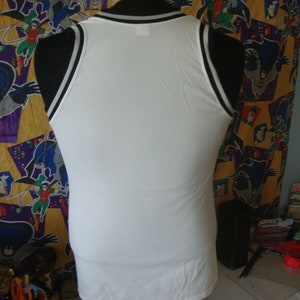 May include: White sleeveless tank top with black and white trim around the neck and armholes. The tank top is on a mannequin against a colorful background.