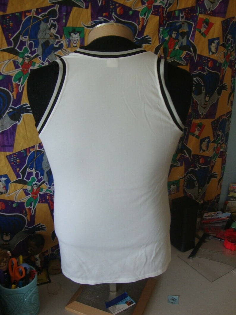 May include: White sleeveless tank top with black and white trim around the neck and armholes. The tank top is on a mannequin against a colorful background.