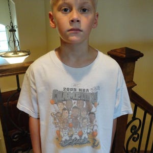 May include: A white t-shirt featuring the text "2005 NBA CHAMPIONS" with a graphic of basketball players. The shirt is worn by a person standing in front of a staircase.