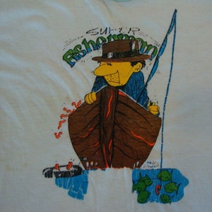 May include: A white t-shirt with a cartoon illustration of a fisherman in a boat. The illustration is in bright colors, including blue, green, orange, and brown. The text "Super Fisherman" is printed above the illustration.