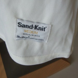 May include: White Sand-Knit t-shirt with a white tag that reads "Sand-Knit" in black text, "Medium" in black text, "Berlin, WI U.S.A." in black text, and "MacGregor Sporting Goods" in black text.