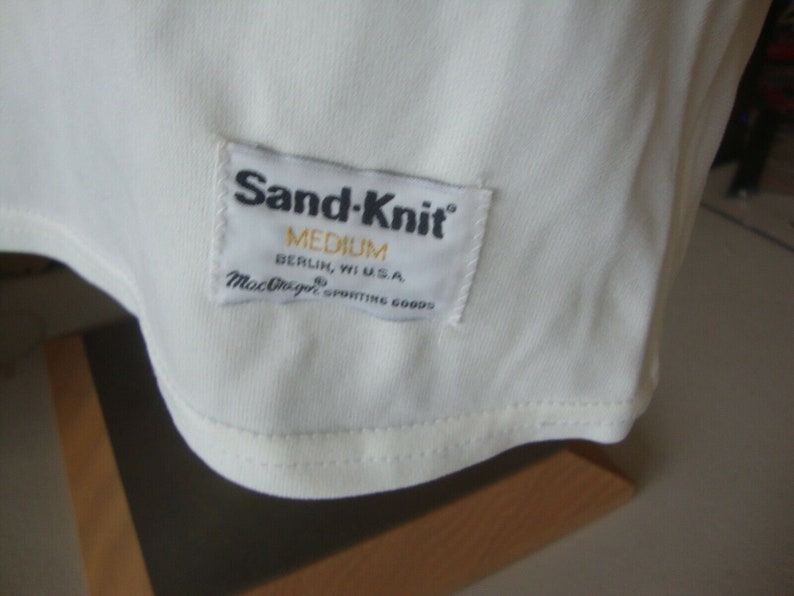 May include: White Sand-Knit t-shirt with a white tag that reads "Sand-Knit" in black text, "Medium" in black text, "Berlin, WI U.S.A." in black text, and "MacGregor Sporting Goods" in black text.