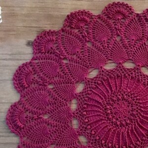 May include: A large, round, crocheted doily in a deep burgundy colour. The doily has a delicate, intricate design with a central motif and radiating patterns.