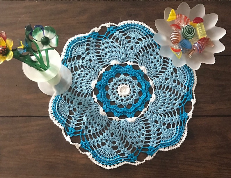 May include: A blue and white crocheted doily with a scalloped edge. The doily is on a wooden table with a vase of flowers and a bowl of sweets.