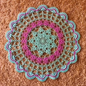 Op de afbeelding: Een gehaakt tafellaken met een delicaat, ingewikkeld ontwerp in tinten turquoise en roze. Het tafellaken heeft een gegolfde rand en een centraal motief.