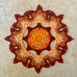 May include: A crocheted doily with a circular design in shades of orange, yellow, brown, and red. The doily has a scalloped edge.