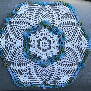 May include: A white crocheted doily with a blue and green border. The doily has a delicate floral design in the center.