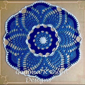 May include: A blue and white crocheted doily with a scalloped edge. The doily has a centre design with a smaller white crocheted circle in the middle. The doily is signed "Gemma R Owen Designs".