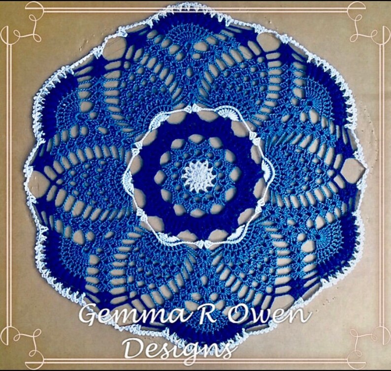 May include: A blue and white crocheted doily with a scalloped edge. The doily has a centre design with a smaller white crocheted circle in the middle. The doily is signed "Gemma R Owen Designs".