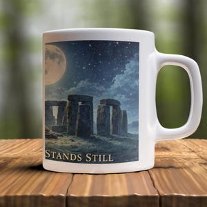 May include: White ceramic mug featuring an image of Stonehenge under a starry night sky with a full moon. The text "STANDS STILL" is printed below the image. The mug has a curved handle and is set on a wooden surface.