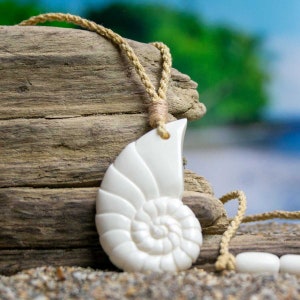 May include: A white nautilus shell pendant necklace with a natural rope cord. The pendant is carved in the shape of a nautilus shell with a spiral design. The necklace is displayed on a piece of driftwood.
