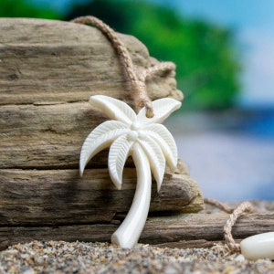 May include: A cream-colored palm tree pendant necklace. The pendant is carved with detailed palm fronds and a curved trunk. The necklace is strung on a natural-colored woven cord. The background is a blurred beach scene.