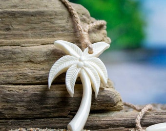 Op de afbeelding: Een crèmekleurige ketting met een palmboomhanger. De hanger is gesneden met gedetailleerde palmbladeren en een gebogen stam. De ketting is geregen aan een natuurlijk gekleurd geweven koord. De achtergrond is een vage strandscène.