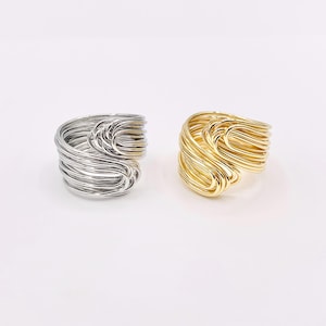 May include: Two rings, one silver and one gold, are displayed against a white background. Each ring is crafted from numerous thin, curved metal wires, creating a woven, sculptural design. The rings are similar in size and shape, showcasing a modern jewelry aesthetic.